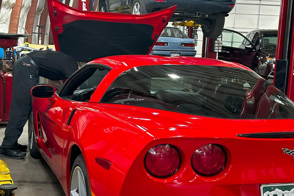 Regular Maintenance near me in Colorado Springs, CO with Legend Motor Works. Image of a red Corvette undergoing regular maintenance with the mechanic inspecting under the open hood at Legend Motor Works.
